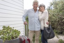 Heureux couple de personnes âgées debout avec valise à l'extérieur de la maison et regardant la caméra — Photo de stock