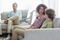 Madre hablando con el hijo en el sofá, mientras que el padre sentado en el fondo en la sala de estar en casa - foto de stock