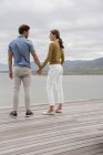 Joven pareja sonriente mirándose y tomándose de la mano en la orilla del lago - foto de stock