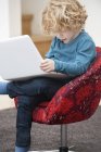 Carino ragazzo con i capelli biondi utilizzando un computer portatile in poltrona a casa — Foto stock
