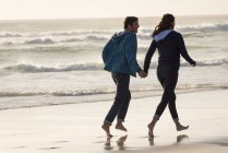 Feliz pareja joven descalza corriendo en la playa al atardecer en otoño - foto de stock