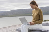 Усміхнена молода жінка використовує ноутбук на березі озера — стокове фото