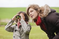 Niño mirando a través de los prismáticos mientras está de pie con la madre en el campo - foto de stock