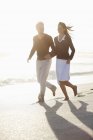 Усміхнена пара бігає на пляжі в сонячному світлі тримаючи руки — стокове фото