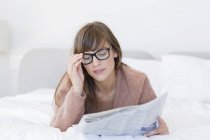 Young woman in eyeglasses reading newspaper in bed — Stock Photo