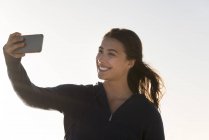 Счастливая женщина делает селфи со смартфоном на фоне ясного неба — стоковое фото