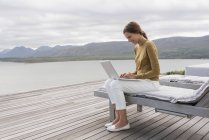 Sonriente mujer joven utilizando el ordenador portátil en la orilla del lago - foto de stock