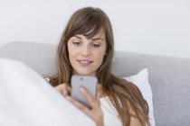 Улыбающаяся молодая женщина общается с мобильным телефоном в постели — стоковое фото