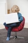 Мальчик с светлыми волосами с помощью ноутбука в кресле дома — стоковое фото