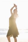 Чуттєва молода жінка в чорній сукні позує на пляжі з піднятими руками — стокове фото