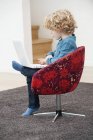 Cute boy with blonde hair using a laptop in armchair at home — Stock Photo