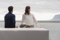Rückansicht eines Paares, das auf einem Hocker am See sitzt und die Aussicht betrachtet — Stockfoto