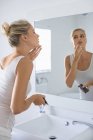 Blonde jeune femme appliquant hydratant sur le visage dans la salle de bain — Photo de stock