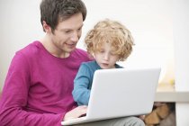 Homem assistindo filho em usar laptop no sofá em casa — Fotografia de Stock