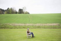 Человек, сидящий на деревянной скамейке и использующий мобильный телефон в зеленом поле — стоковое фото