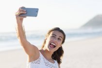 Игривая молодая женщина делает селфи со смартфоном на пляже — стоковое фото