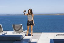 Stylish young woman taking selfie with smartphone on terrace at lake shore — Stock Photo
