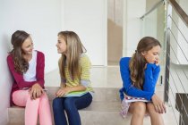 Teenage girl sitting on stairs and thinking while female friends whispering — Stock Photo