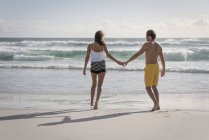 Вид сзади на молодую пару, идущую по пляжу, держась за руки — стоковое фото