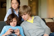 Menino usando um celular com seu irmão e irmã em casa — Fotografia de Stock
