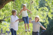 Frères et sœurs souriants jouant dans le swing des arbres dans le jardin d'été — Photo de stock