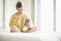 Woman in yellow fluffy sweater using phone and eating chocolate on bed — Stock Photo