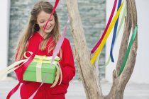 Усміхнена дівчина тримає подарункову коробку біля дерева з різнокольоровими стрічками — стокове фото