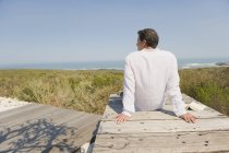 Rückansicht eines Mannes, der auf der Strandpromenade an der Küste sitzt und die Aussicht betrachtet — Stockfoto