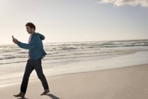 Young man using mobile phone and earphones while walking on beach — Stock Photo