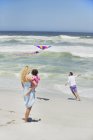 Mutter trägt Kind, während Mann Drachen am Strand fliegen lässt — Stockfoto