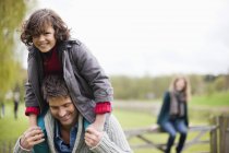 Hombre llevando sonriente hijo en hombros al aire libre con la madre en el fondo - foto de stock