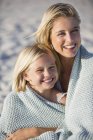 Крупный план улыбающейся блондинки и дочери, сидящих на песчаном пляже, завернутых в полотенце — стоковое фото
