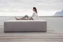 Happy mature woman sitting on ottoman at lake shore and looking away — Stock Photo
