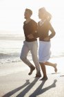 Smiling couple running on beach in sunlight holding hands — Stock Photo
