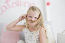 Portrait of little girl making mustache with hair on bed — Stock Photo