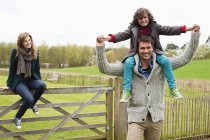 Man carrying son on shoulders while mother sitting on wooden fence in countryside — Stock Photo