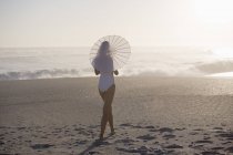 Mujer delgada en traje de baño caminando en la playa con sombrilla - foto de stock
