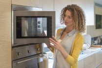 Женщина с вьющимися волосами смотрит в микроволновку на кухне — стоковое фото