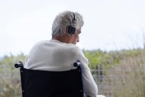 Старший чоловік у інвалідному візку слухає музику з навушниками на відкритому повітрі — стокове фото