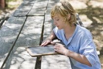 Мальчик-подросток пишет на доске за деревянным столом на открытом воздухе — стоковое фото