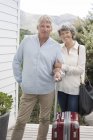Happy senior couple standing with suitcase outside of house and looking at camera — Stock Photo