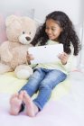 Happy little girl using digital tablet with teddy bear on bed — Stock Photo