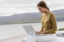 Smiling young woman using laptop on lake shore — Stock Photo