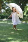 Cute little girl in white summer dress holding umbrella in sunny garden — Stock Photo