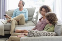 Madre che parla al figlio sul divano mentre il padre guarda la tv sullo sfondo in soggiorno a casa — Foto stock