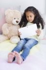 Little girl using digital tablet with teddy bear on bed — Stock Photo