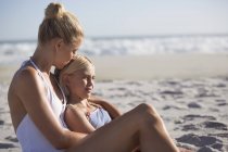 Mujer feliz y su hija sentados en la playa soleada - foto de stock