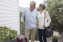 Happy senior couple standing with suitcase outside of house — Stock Photo
