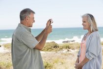 Чоловік фотографує дружину з мобільним телефоном на пляжі — стокове фото