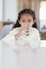 Маленькая девочка пьет воду из стекла за столом дома и смотрит в сторону — стоковое фото
